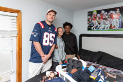 Three people standing in a bedroom with a New England Patriots jersey, casual clothing, and a baseball cap. The bed has a sports-themed comforter and a laundry basket with household items. A football game poster is on the wall.