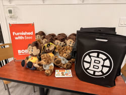 Red wooden table with plush toys including brown bears and monkey mascots wearing yellow and gray shirts, next to black reusable bags with a white "B" logo. A red sign reads "Furnished with love BOB Discount Furni." The setup promotes Bob's Discount Furniture branded merchandise.