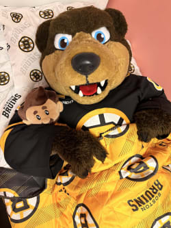 Boston Bruins plush mascot wearing a black jersey and covered with a yellow Bruins blanket on a bed with matching Boston Bruins pillowcases.