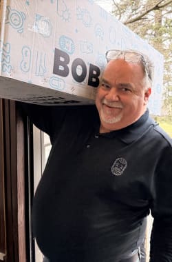 Man wearing black polo shirt with a bearded face logo carrying a large white and blue box labeled "BOB" on his shoulder, standing in a doorway.