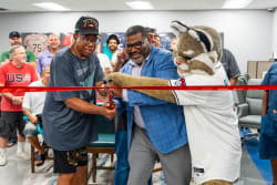 A ribbon-cutting ceremony with a man in a blue blazer and glasses, a man in a black USA Veterans cap and casual clothes, and a mascot in a white sports jersey cutting the red ribbon. Several people watch in the background.