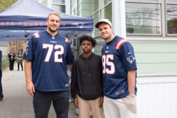 Two men wearing navy blue New England Patriots NFL jerseys with numbers 72 and 85 stand on either side of a young person in a black hoodie and beige pants, posing outdoors near a tent and a green building.