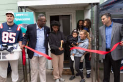 A diverse group of people participates in a ribbon-cutting ceremony outside a building, with a young person holding large gold scissors cutting a red ribbon. A man in a Patriots jersey holds a basket of items, and a blue sign in the background reads "BOB'S Discount Furniture Welcome." The scene highlights a community event promoting local business and family involvement.
