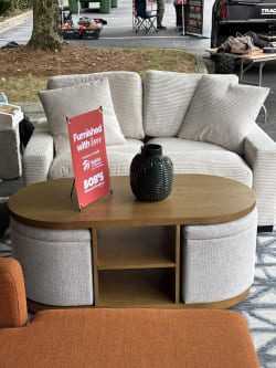 Beige corduroy loveseat with matching pillows behind a wooden oval coffee table featuring fabric-covered storage compartments and a black ceramic vase. A red sign on the table reads: "Furnished with love. Donation to Benefit Habitat for Humanity of Greater Atlanta. BOB'S Discount Furniture."