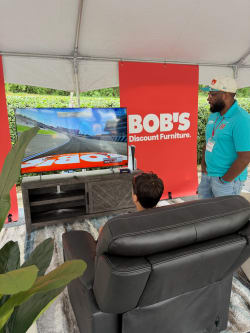 A man in a turquoise shirt and cap watches a racing video game on a flat-screen TV, displayed on a wooden console. A child sits on a black leather recliner. Behind the TV is a red banner with white text reading "BOB'S Discount Furniture." The setup is under a white tent with a gray and white rug underneath.