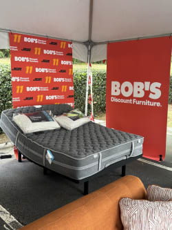 Adjustable gray mattress on black metal frame with two pillows under a tent, displayed with red banners reading "BOB'S Discount Furniture" for outdoor furniture sale.
