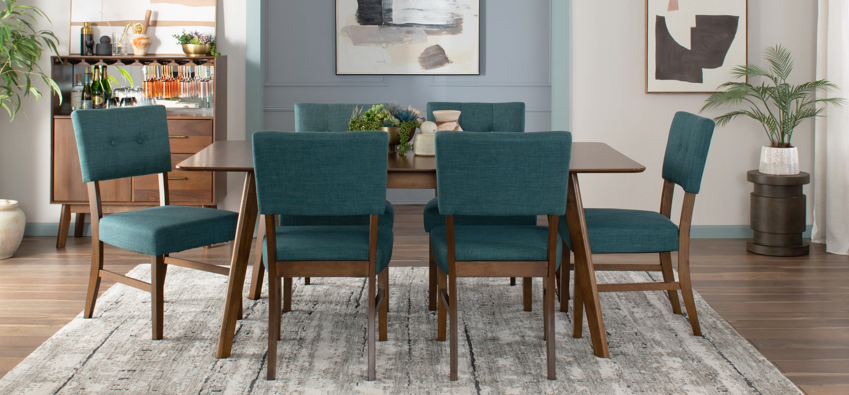 Dining room set with six teal upholstered chairs featuring wooden legs and a rectangular wooden table on a gray patterned rug.