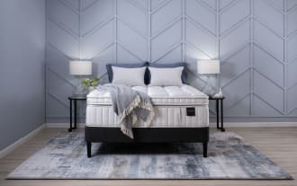 Obsession 16.5'' King Ultra Plush Mattress with white quilted top, plush layers, graphite-infused memory foam, and individually wrapped coils on black base, styled with gray pillows and throw blanket in modern blue-gray bedroom.