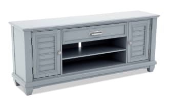 Gray wooden TV stand with two shutter-style cabinet doors, a central drawer with metal handle, and open shelving with cable management hole.