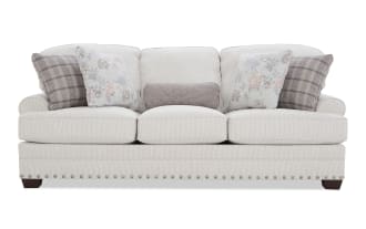 Cream-colored corduroy fabric sofa with rolled arms, three seat cushions, nailhead trim along the base, and five decorative pillows in floral and plaid patterns.