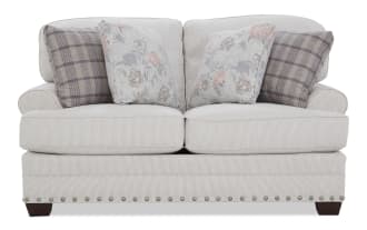 Two-seater beige fabric loveseat with subtle pinstripes, featuring four decorative cushions in floral and plaid patterns, wooden legs, and nailhead trim.