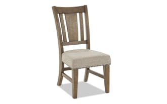 Wooden dining chair with beige upholstered seat, nailhead trim, and slatted backrest, featuring a rustic finish and sturdy legs.