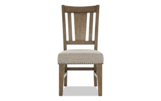 Wooden dining chair with beige upholstered seat, nailhead trim, and slatted backrest, featuring a rustic finish and sturdy legs.