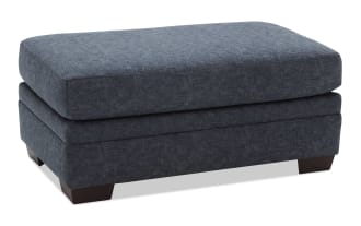 Dark blue fabric upholstered rectangular ottoman with cushioned top and wooden block legs, suitable for living room seating or footrest.