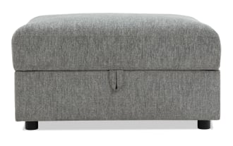 Gray fabric upholstered storage ottoman with a cushioned top, rectangular shape, and black cylindrical legs for living room seating and storage.