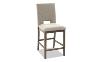 Upholstered beige fabric bar stool with cushioned seat, wooden frame, and rectangular cutout in backrest, modern dining chair design.