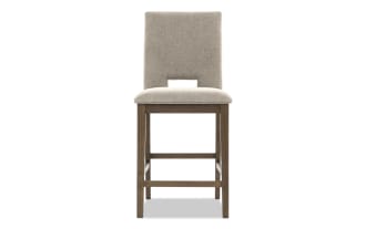 Upholstered beige fabric bar stool with cushioned seat, wooden frame, and rectangular cutout in backrest, modern dining chair design.