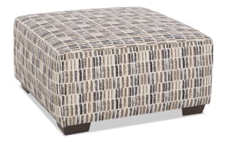 Rectangular upholstered ottoman with a textured fabric cover featuring a vertical striped pattern in beige, brown, and black tones. The ottoman has dark wooden block legs and a cushioned top, suitable for use as a footrest or extra seating in living room decor.