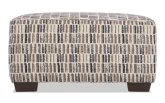 Rectangular upholstered ottoman with a textured fabric cover featuring a vertical striped pattern in beige, brown, and black tones. The ottoman has dark wooden block legs and a cushioned top, suitable for use as a footrest or extra seating in living room decor.