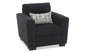 Dark gray upholstered armchair with wide padded arms, cushioned back, and a rectangular patterned lumbar pillow, featuring wooden block legs.