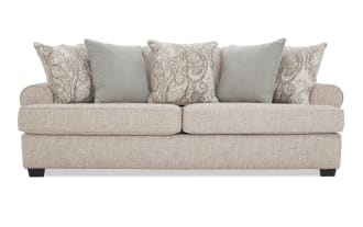 Beige fabric sofa with rolled arms, two large seat cushions, and five decorative throw pillows in paisley and solid patterns, modern style.