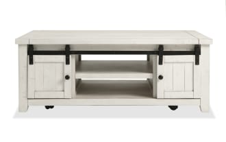 Rustic Montana White Storage Coffee Table from Montana Entertainment & Occasional collection, made of pine solids and plywood, with white finish, gliding barn doors, black metal hardware, open shelving, and casters for easy mobility.