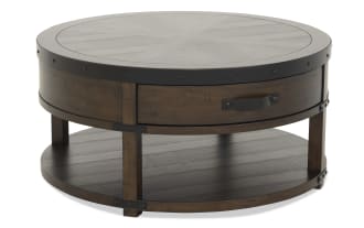 Round wooden coffee table with dark brown finish, metal rivet accents, single drawer with black handle, and lower shelf for storage.