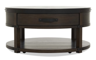 Round wooden coffee table with dark brown finish, metal rivet accents, single drawer with black handle, and lower shelf for storage.