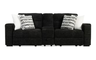 Black fabric reclining loveseat with wide armrests, two patterned gray and white accent pillows, and two white fluffy pillows, featuring a central console with storage.