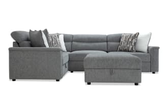 Gray fabric sectional sofa with matching ottoman, featuring plush cushions, patterned and solid throw pillows, and a modern design.