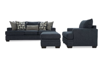 Dark blue fabric sofa set with three seat cushions, matching ottoman, and patterned throw pillows, modern living room furniture.