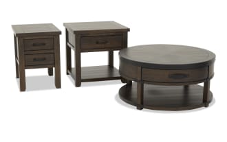 Set of three dark brown wooden living room tables including a round coffee table with metal accents and two matching end tables with drawers.