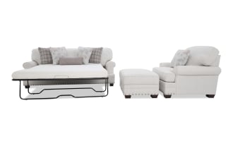Beige fabric sleeper sofa with floral and plaid throw pillows, paired with matching armchair and ottoman featuring nailhead trim and wooden legs.