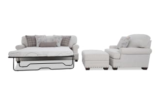 Beige fabric sofa set with floral and plaid throw pillows, featuring a pull-out bed frame and matching ottoman with nailhead trim.