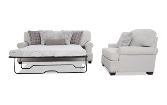 Beige fabric sleeper sofa with floral and plaid throw pillows, metal frame fold-out mattress, and nailhead trim on the base.