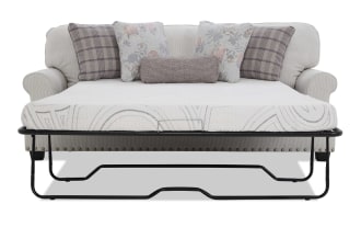 Light beige fabric sofa bed with rolled arms, nailhead trim, and black metal frame. Includes white quilted mattress and decorative floral and plaid pillows.