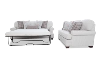 Light beige fabric sleeper sofa with rolled arms, dark wooden legs, and nailhead trim; includes floral and plaid throw pillows and a pull-out mattress.