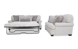 Light beige fabric sofa with rolled arms and nailhead trim, shown with floral and plaid throw pillows and a metal frame pull-out sleeper mattress extended.