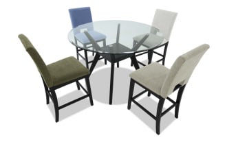 Round glass dining table with black wooden base and four upholstered chairs in blue, green, beige, and off-white fabric.