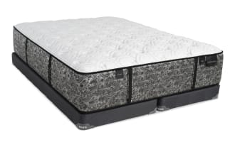 Mattress with a white quilted top and gray paisley-patterned sides labeled "PLUSH," "FIRM," and "DISTINCTION," resting on a dark gray Bob's mattress foundation.