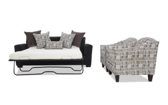 Black fabric sleeper sofa with metal frame mattress extended, decorated with patterned and solid throw pillows, next to two matching patterned armchairs with dark wooden legs.