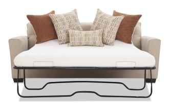 Beige fabric sofa bed with a white mattress and metal frame, featuring four decorative pillows in rust and patterned beige tones, ideal for small spaces.