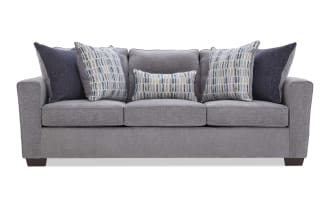 Modern gray fabric sofa with three seat cushions, wide armrests, and five decorative pillows in blue and patterned designs, ideal for living room seating.