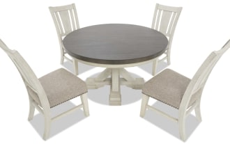 White wooden dining table set with beige upholstered cushioned chairs, featuring nailhead trim and a round pedestal table top.