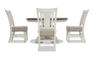White wooden dining table set with beige upholstered cushioned chairs, featuring nailhead trim and a round pedestal table top.