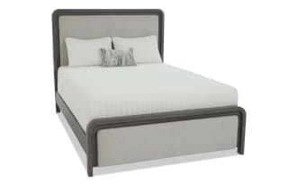 Queen-size bed with dark wood frame and light beige upholstered headboard and footboard, featuring white quilted bedding and decorative pillows.