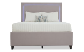 Modern upholstered bed with a light mauve fabric frame and headboard featuring built-in soft blue LED lighting, styled with white quilted bedding and decorative pillows.