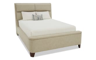 Beige upholstered bed with padded headboard and footboard, white quilted bedding, decorative blue and beige accent pillow, wooden legs, modern style.