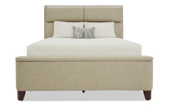Beige upholstered bed with padded headboard and footboard, white quilted bedding, decorative blue and beige accent pillow, wooden legs, modern style.