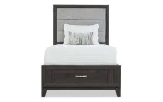 Twin bed with dark wood frame and upholstered gray fabric headboard, featuring a front drawer for storage and white bedding with decorative pillows.